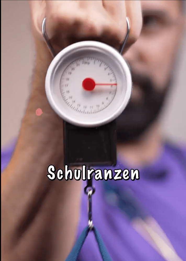 Nahaufnahme: Hand hält eine Federwaage mit Zeiger, daran hängt ein Schulranzen, Schriftzug Schulranzen