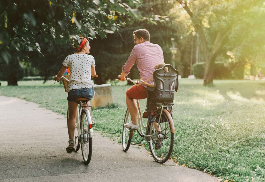 Foto: Zwei Erwachsene radeln durch einen sonnigen Park, am hinteren Fahrrad ist ein Kindersitz montiert