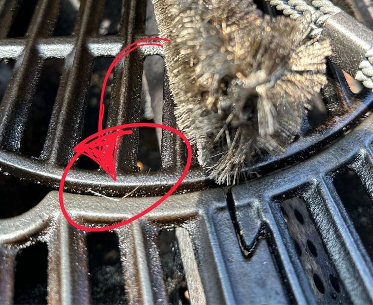 Foto: Nahaufnahme eines Grillrosts mit Drahtbürste, ein roter Pfeil markiert eine gelöste Drahtborste